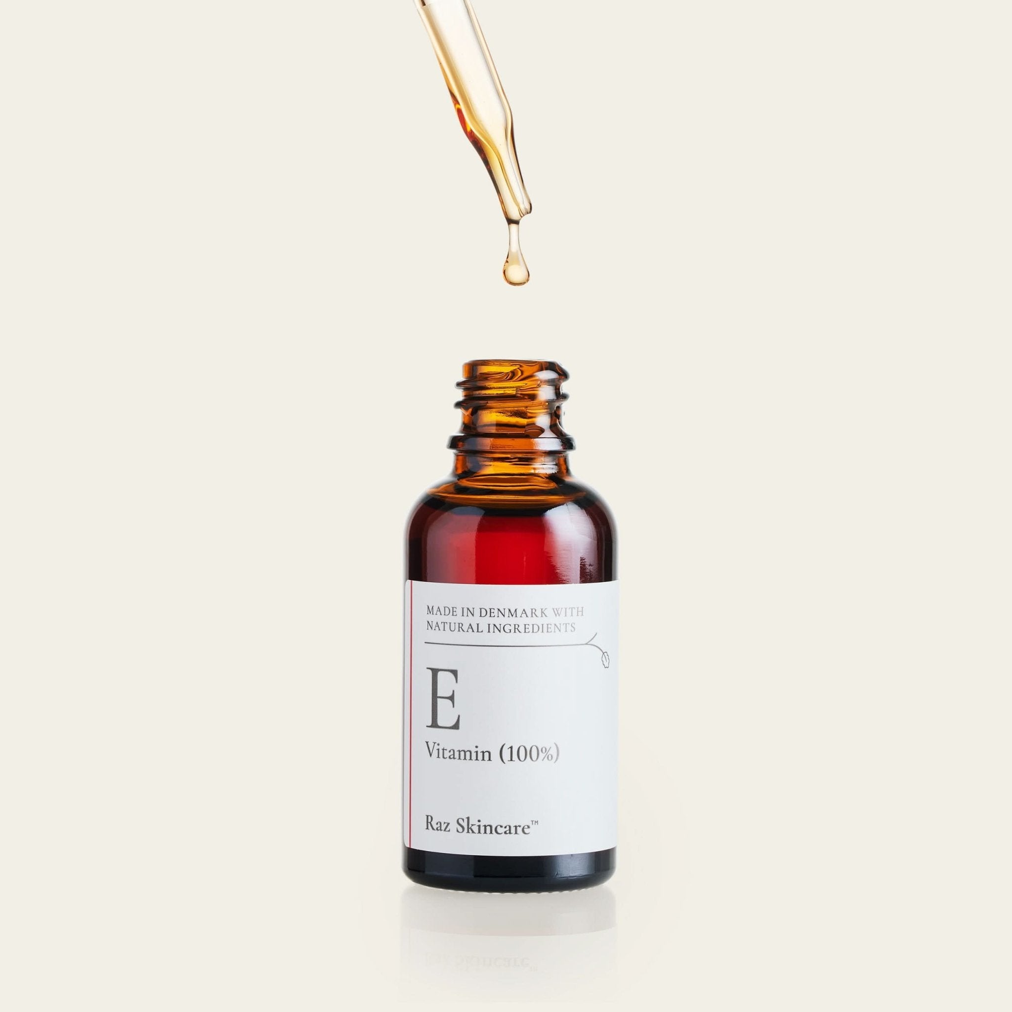 En pipette taget op fra e-vitamin flasken og holdes ovenover mens en dråbe bliver pumpet ud, placeret foran en beige baggrund