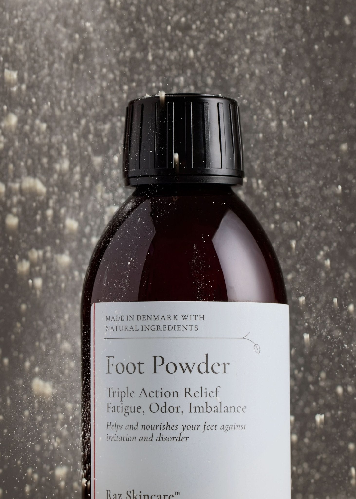 Foot Powder 75g - Raz Skincare - Raz Skincare Foot Powder i brun flaske med sort låg, omgivet af dryssende pulverpartikler